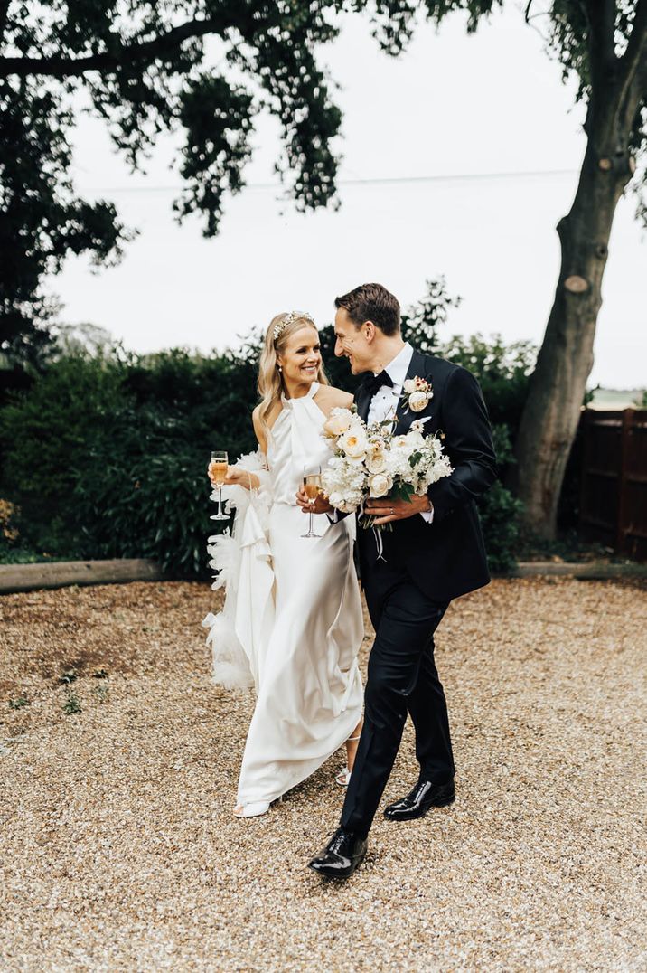 Bride in halter neck wedding dress with feather accessories walking hand in hand with the groom in black tie holding the bride's neutral bouquet