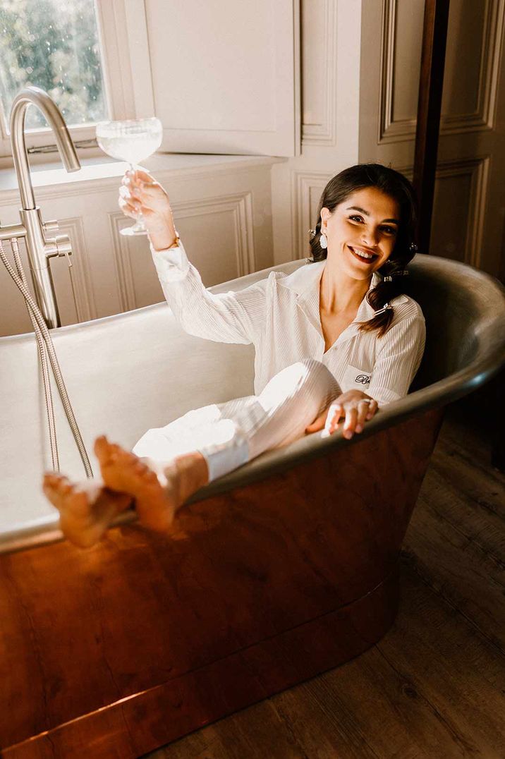 Bride in white satin bridal pyjamas having a glass of wine in the bath at Chapel House Estate wedding venue