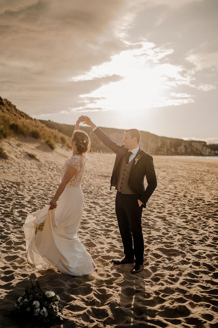 Groom in three piece suit spins the bride around at the beach on their wedding day 