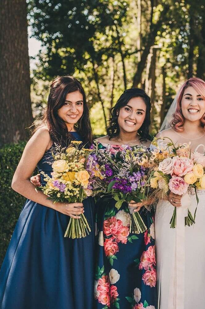 Bridesmaids wearing assorted short navy bridesmaid dresses with floral detail and off shoulder sleeves. They are holding huge bouquets of bright spring flowers | Claire Penn Photography