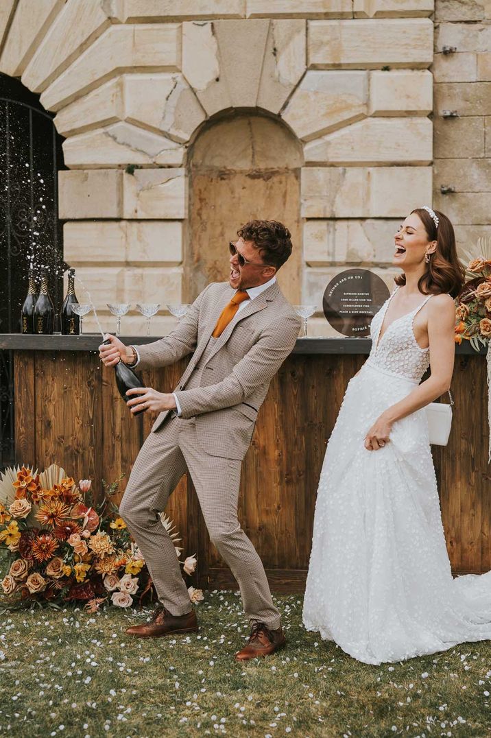 Couple in sleeveless lace wedding dress and tartan suit doing champagne pop with white confetti