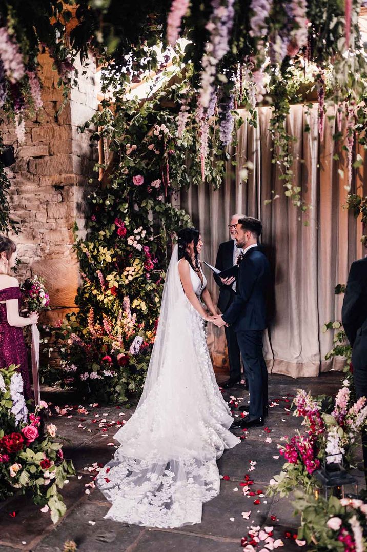 Bride in v neck sleeveless wedding dress with church length embroidered sheer lace veil standing with groom in classic black tie surrounded by garden rose, foliage, and dried flower floral arrangements at Cripps Barn wedding venue 