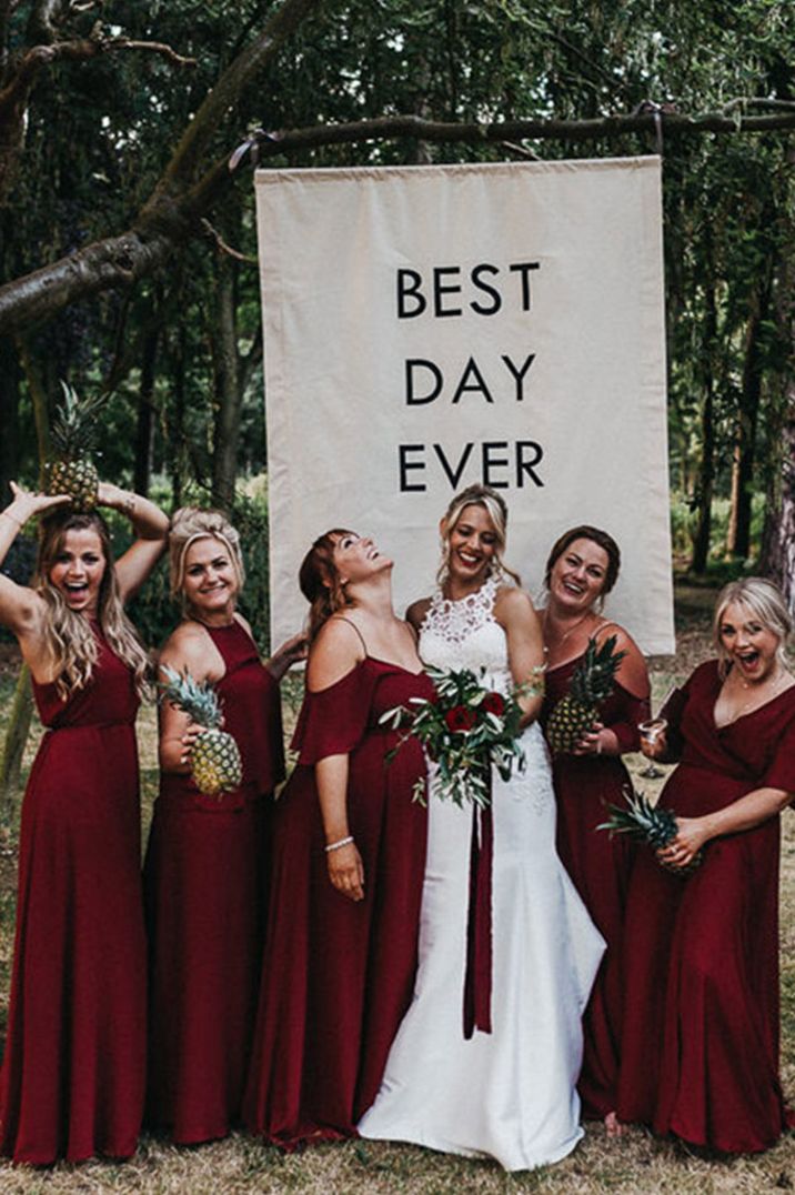 Bridesmaids in deep red wine coloured bridesmaid dresses with the bride in front of wedding banner sign 