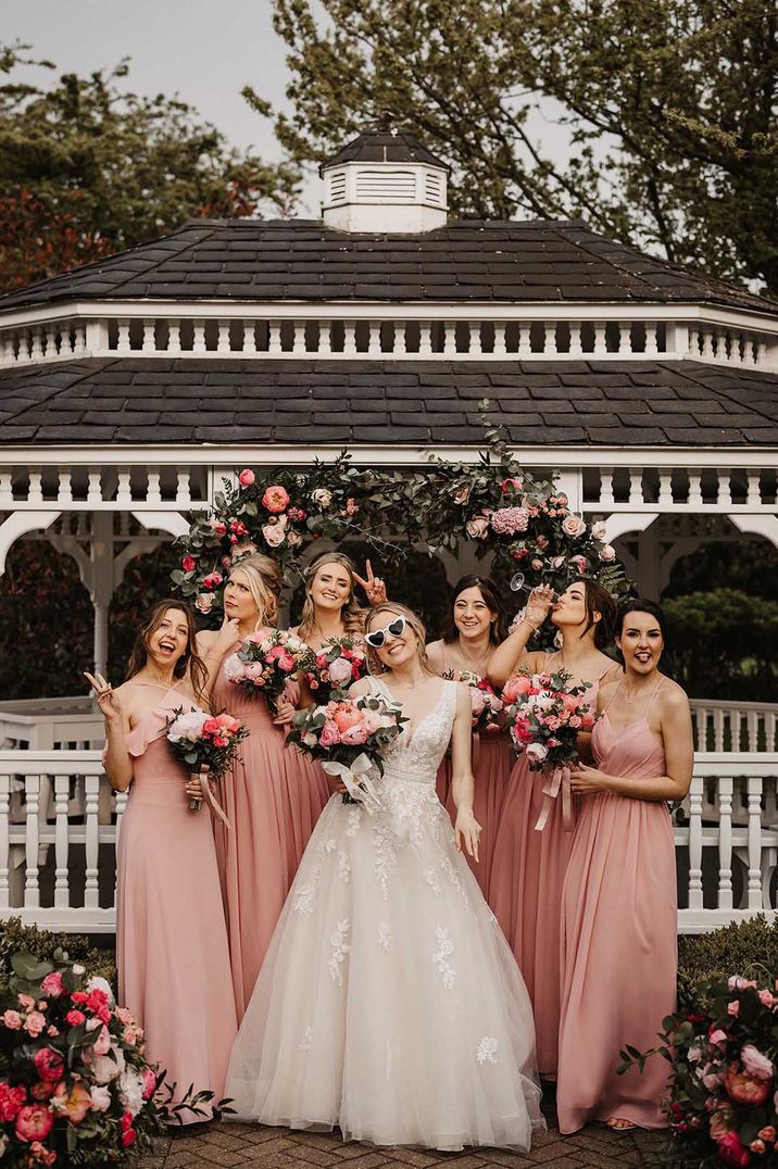 Bride in 3d applique embellished wedding dress standing by pink flower arch in wedding sunglasses with the bridal party in sleeveless pink bridesmaid dresses 