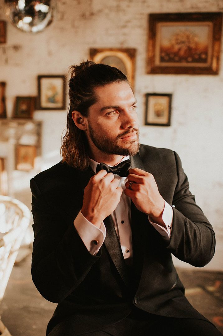 A groom with long hair in a half up half down style tightens his bowtie.