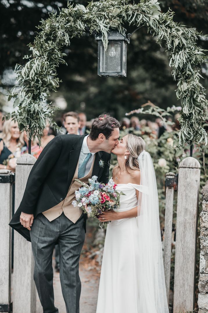 Traditional church wedding with groom in a morning suit and bride in an off the shoulder wedding dress kissing at the gate of the church