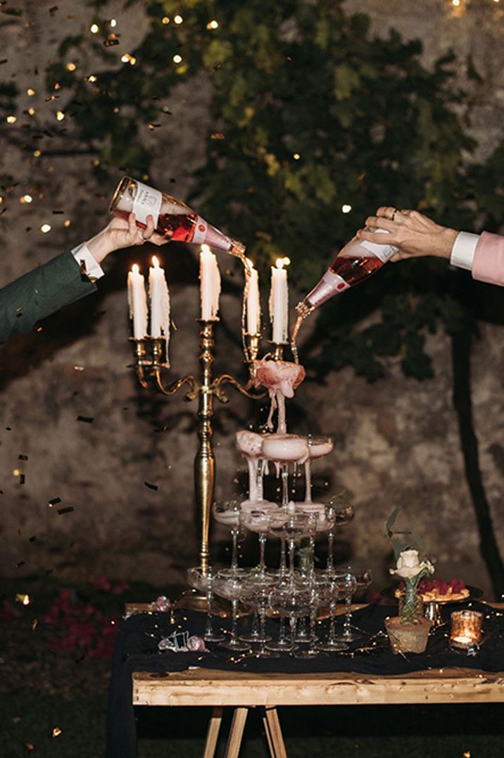 Two grooms pouring pink champagne into a champagne tower at candlelight 