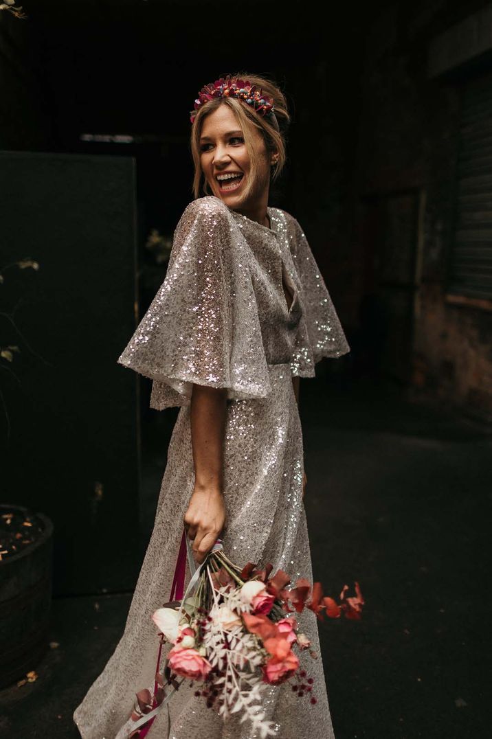 Bride wears sparkly wedding dress with short flowing sleeves and split to front whilst carrying colourful floral bouquet 