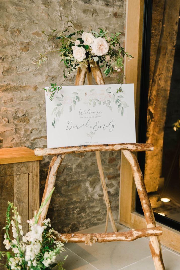 White welcome wedding sign on wooden easel with rose and foliage decorations at barn wedding venue