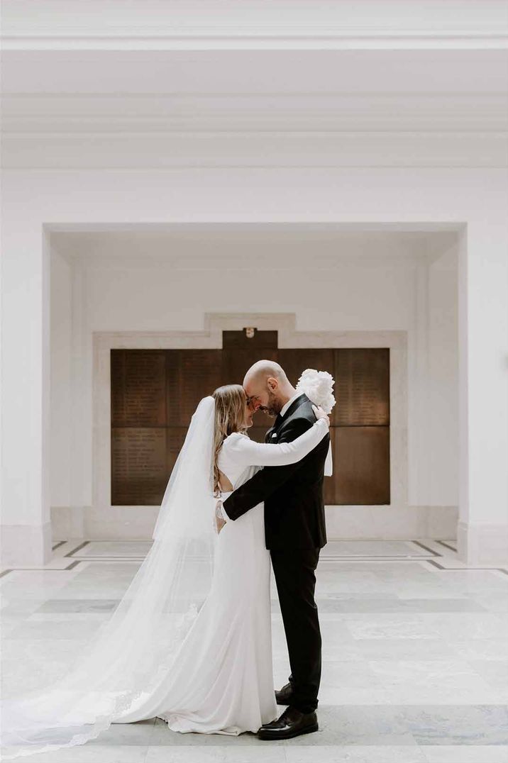 Bride in long sleeve wedding dress with church-length veil embracing groom in classic black tuxedo in all-white reception room of Shoreditch Studios London 