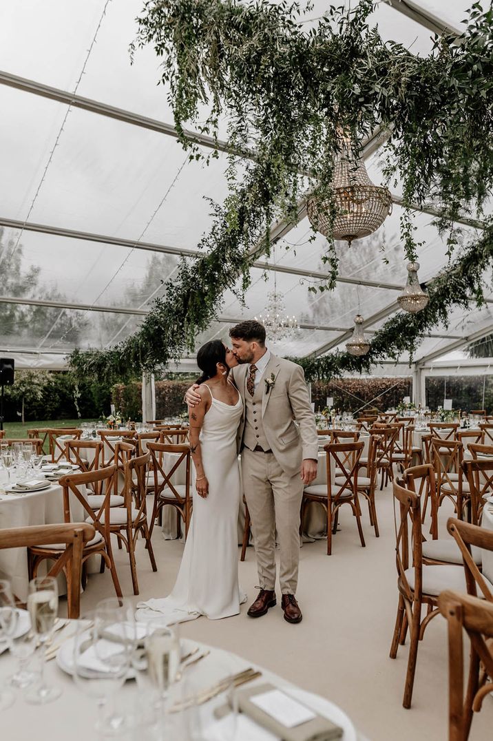 Bride in cowl neck wedding dress at glass marquee wedding with chandelier and foliage decor with the groom in grey suit 