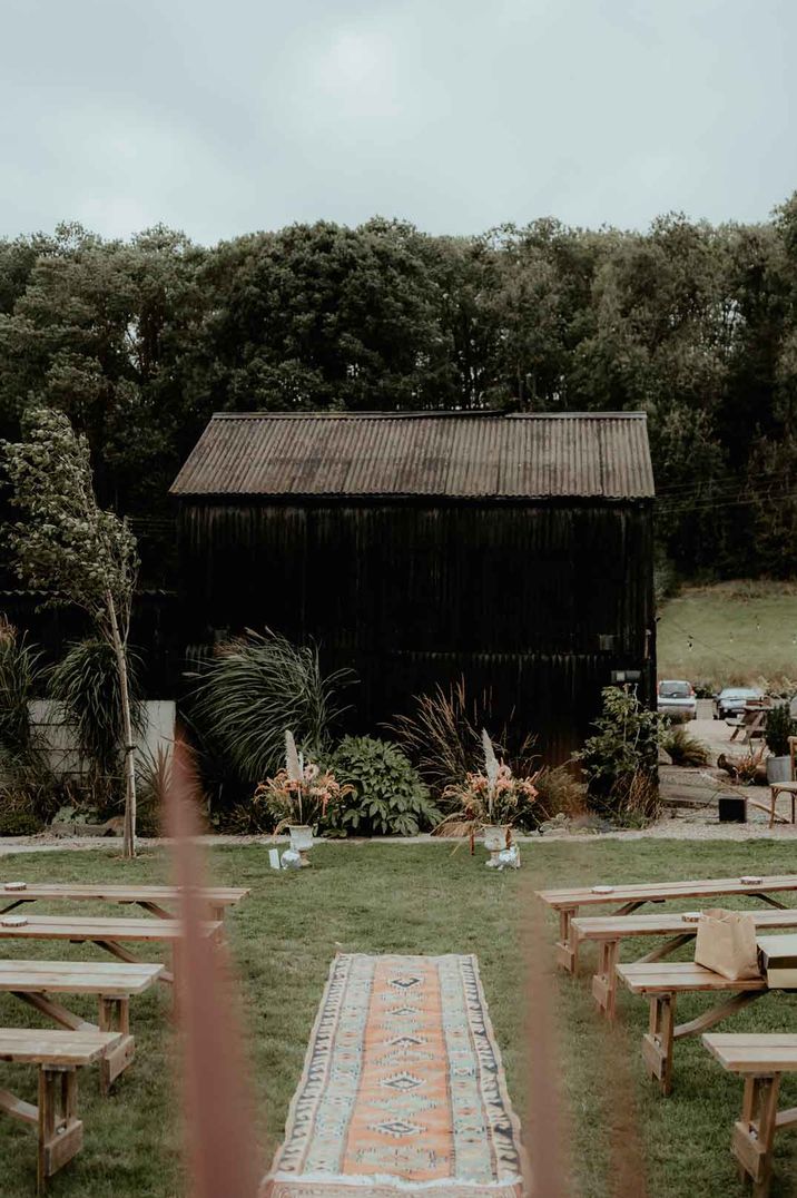 Rustic boho wedding alter at Rhyse Farm wedding venue with rug going down the aisle, wooden bench seats and dried flowers and plants blowing in the wind 