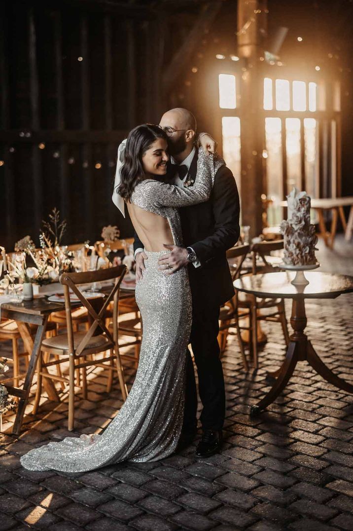 Bride in silver sparkly wedding dress with open back detail dancing with groom in classic black tuxedo in front of rustic luxe wedding tablescape and decor at Elmley Nature Reserve wedding venue