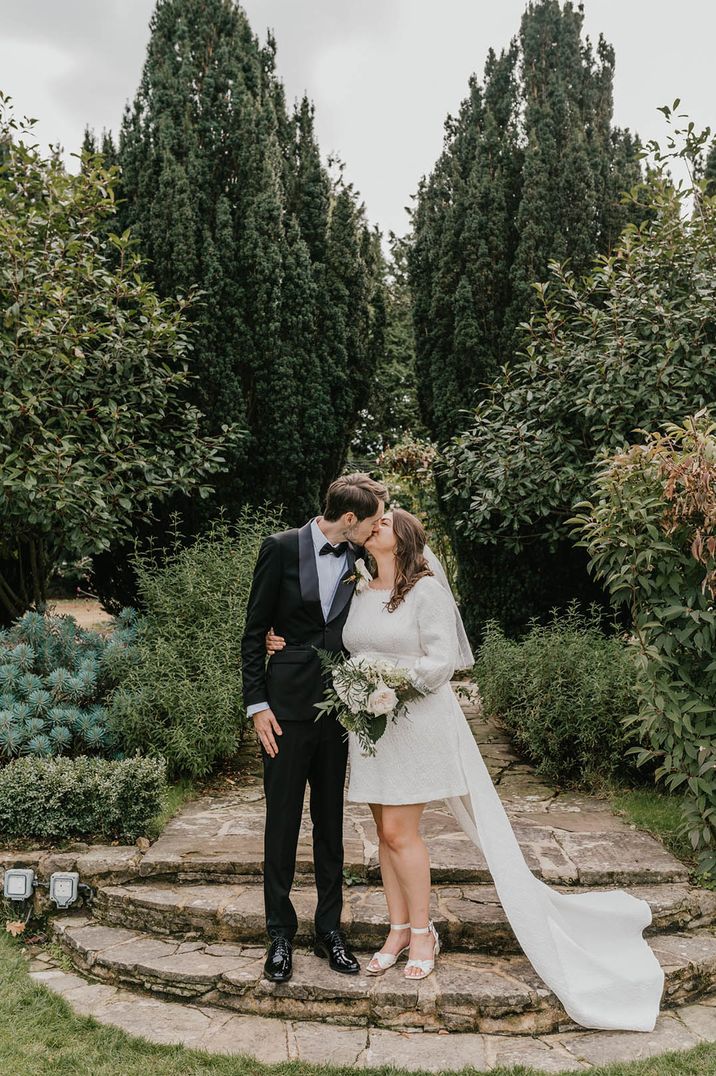 Bride wearing long sleeve short wedding dress with embellishments with long train kissing the groom