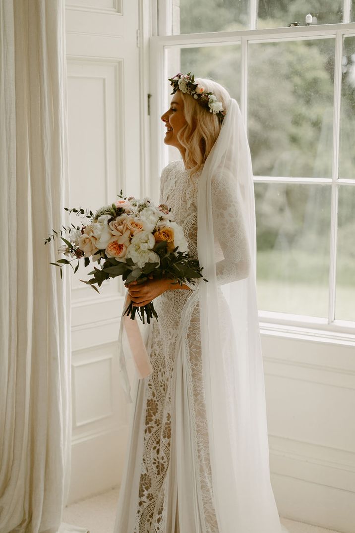 Bride in lace wedding dress holding wedding bouquet standing in front of window with white curtains