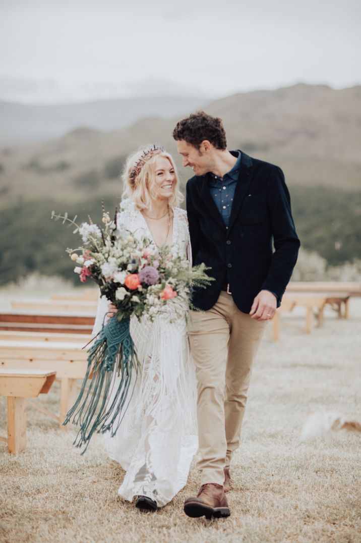 Bride in a boho fringe wedding dress holding a wildflower bouquet with macrame ribbon laughing with her husband in beige chino and a navy blue blazer