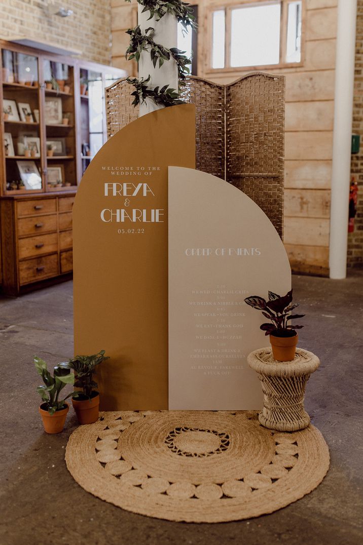 Wedding welcome sign and oder of events notice board in deep mustard and cream with potted plants and wicker details