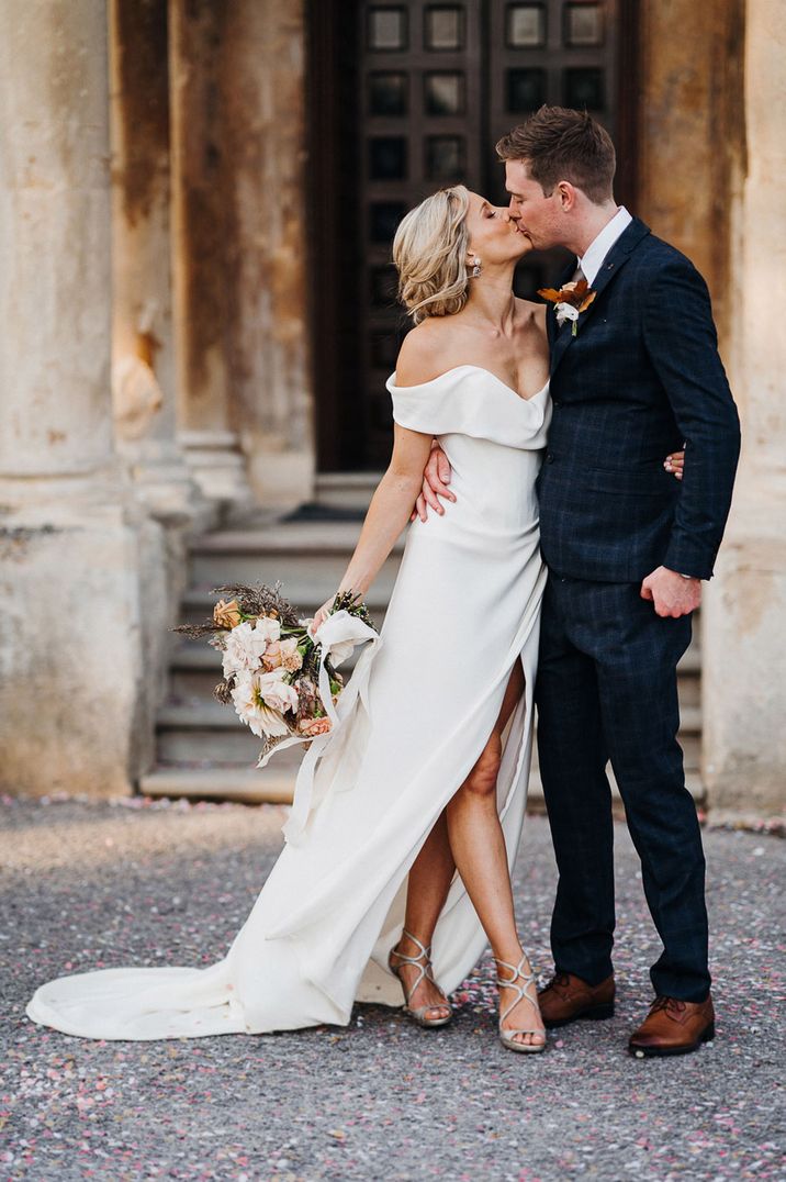 Groom in blue checkered suit and bride in off the shoulder wedding dress and silver heels share a kiss