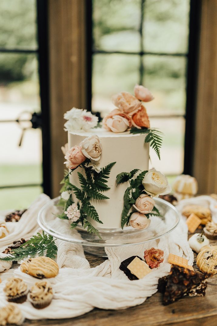 Single tier wedding cake on a glass cake stand decorated with flowers and fern leaves