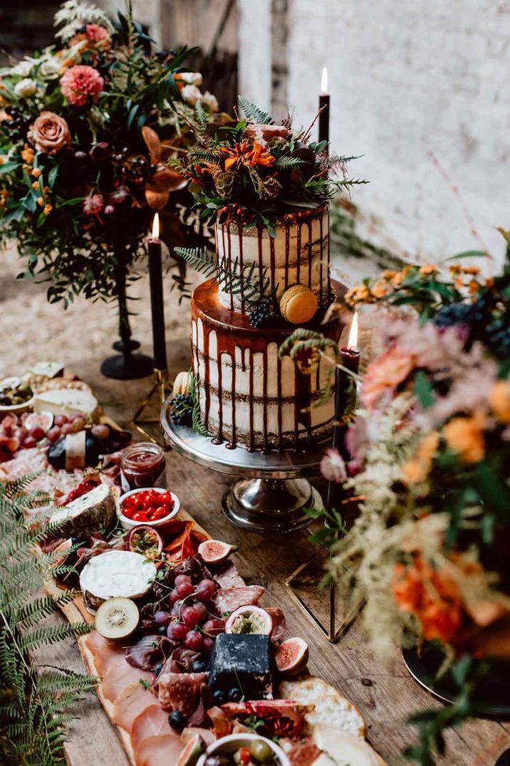 Two tier buttercream wedding cake with chocolate drip icing and dried flower decorations with dark tapered candles and a platter of fruit 