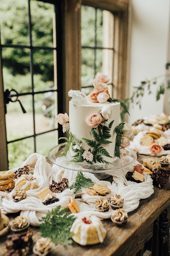 Dessert table covered in yummy desserts with the single tier tall white iced wedding cake decorated with pink and white peonies with fern leaves 