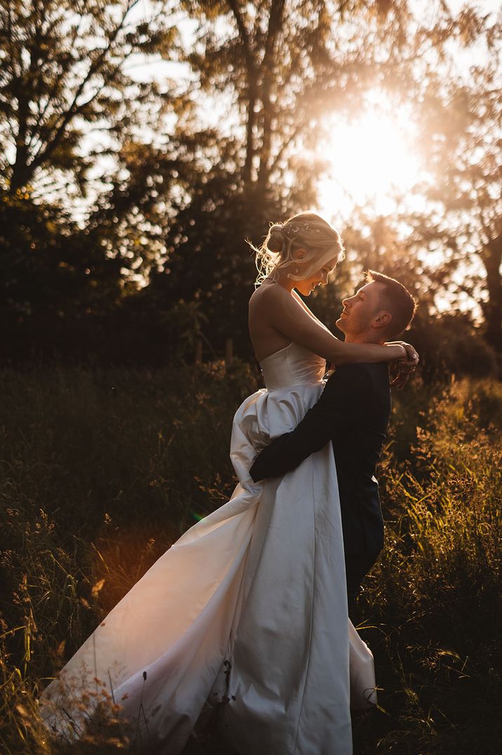 Bride wearing her hair in a blonde updo in a strapless wedding dress being lifted up by the groom as the sun shines through the trees