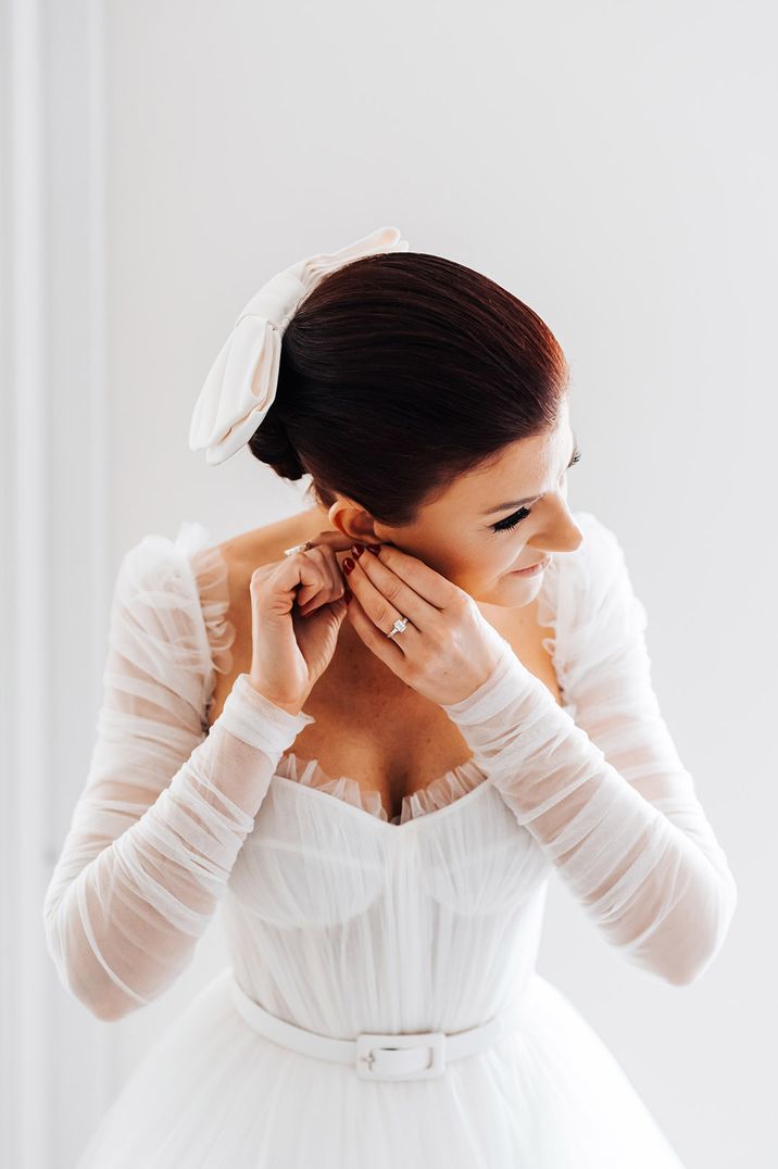 bride in a long sleeve Emma Beaumont wedding dress putting her earrings on