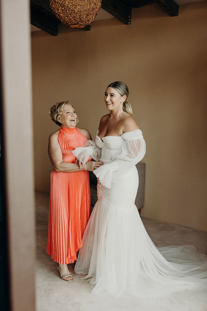 Bride in off the shoulder mermaid wedding dress with mother of the bride in a bright orange halter neck satin dress 