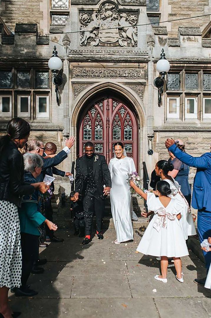 Pregnant bride wearing a minimalist long sleeve satin gown and groom exiting the registry office with guests throwing confetti