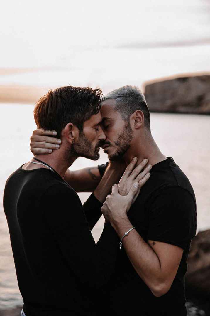 Two men embrace each other at their beach engagement shoot and lean in for a kiss