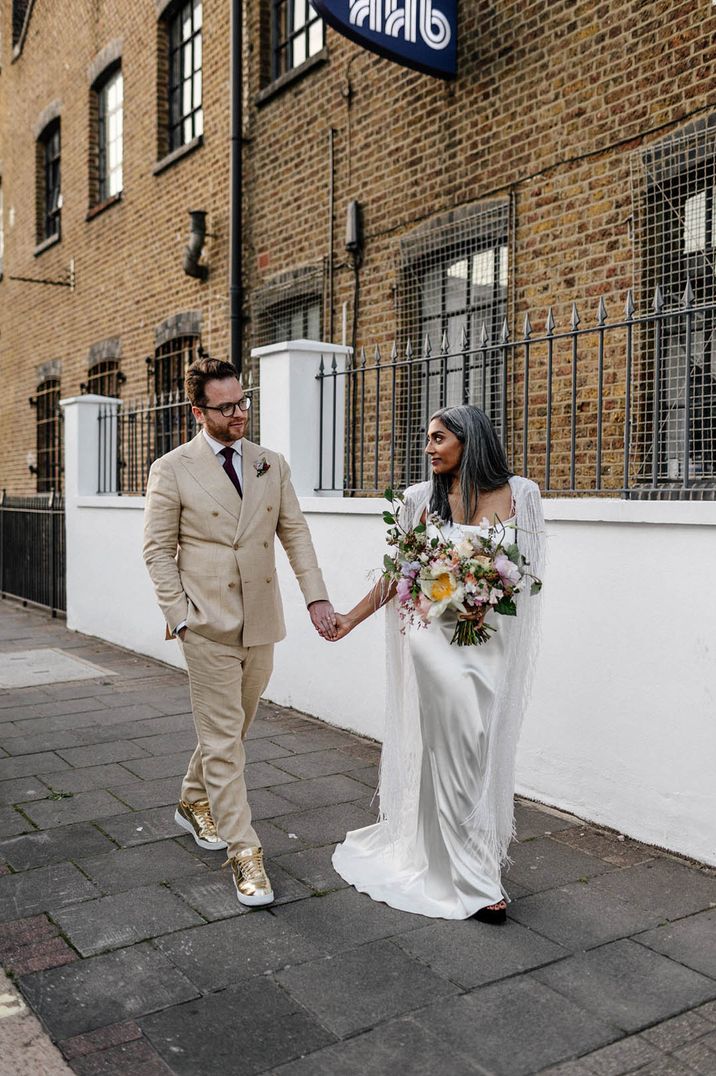 Groom in double breasted beige wedding suit with shiny gold shoes walking with the bride in a silver tassel bridal cape 