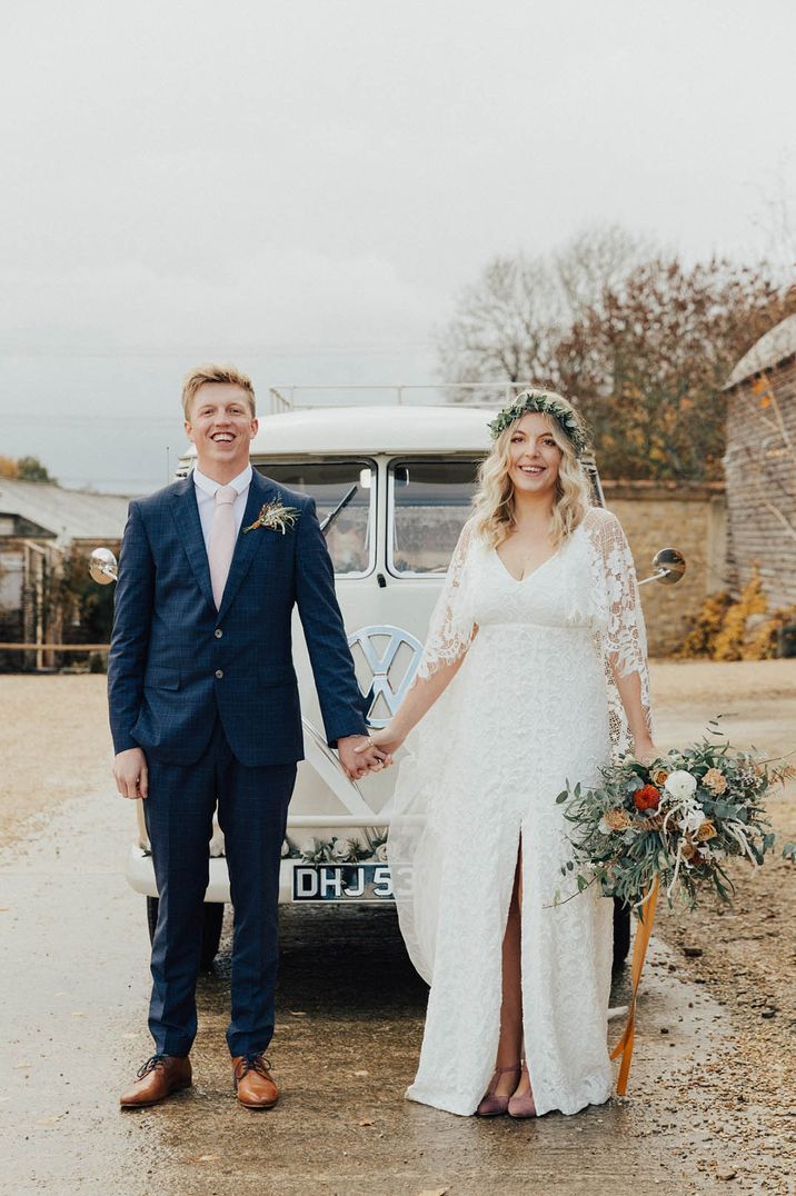 Bride in long sleeve floral lace wedding dress and floral crown and groom in dark blue suit in front of classic VW cream camper van