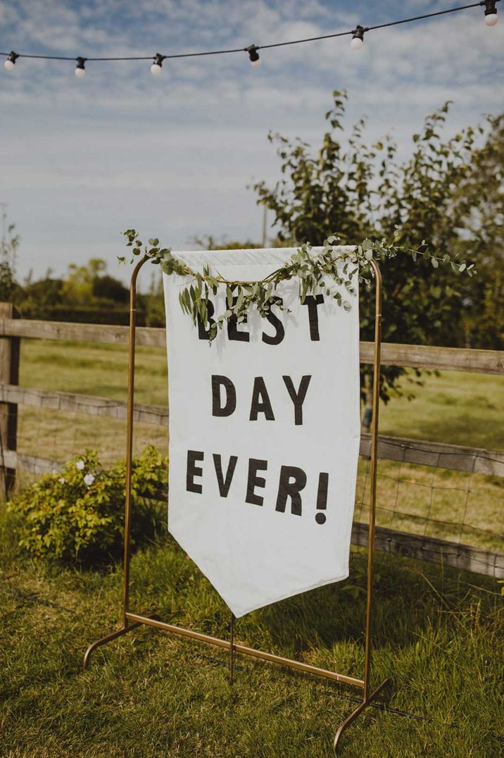 DIY white fabric wedding sign reading "Best Day Ever!" on copper sign stand with floral details around the sides