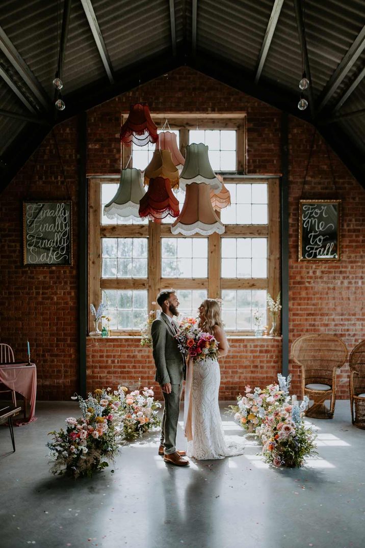 Bride in sleeveless lace wedding dress holding large colourful rose bouquet with foliage and groom in grey suit standing in Giraffe Shed wedding venue with lots of colourful rose floral decorations on the floor and six textured lampshades in sage green, dark red, pink and burnt orange