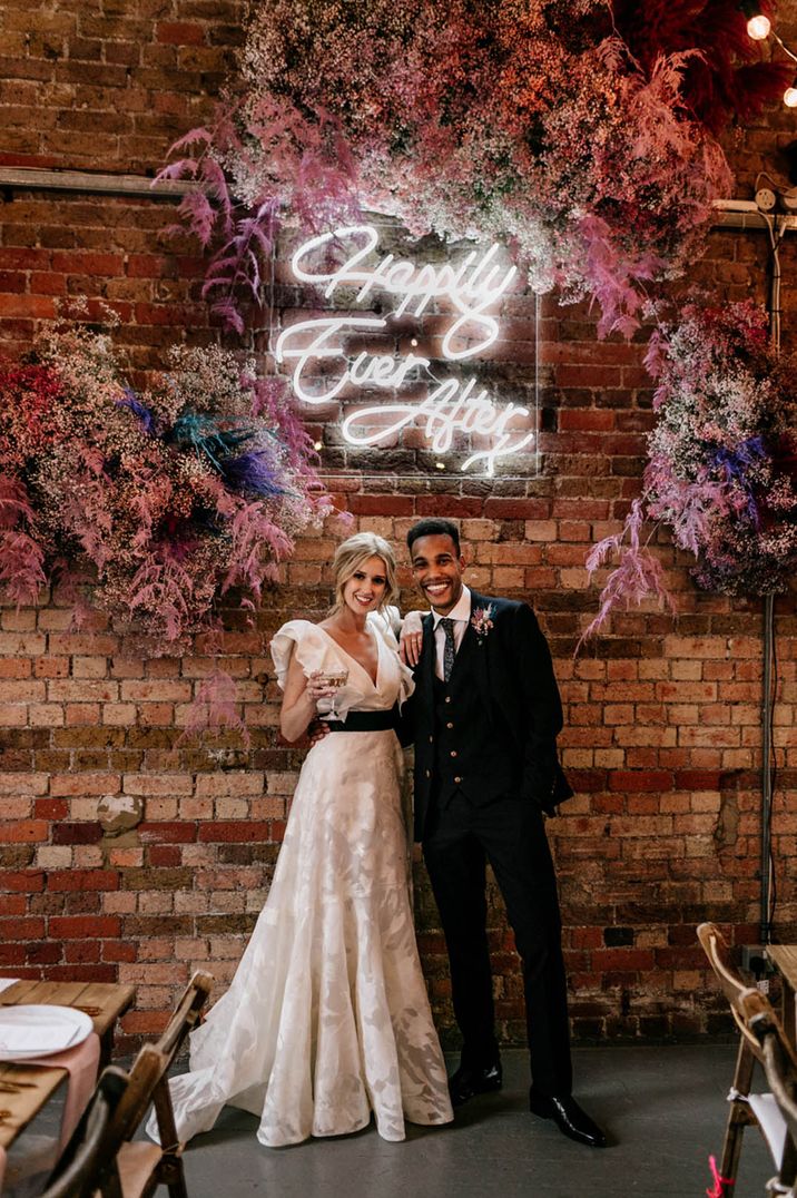 Bride in a ruffle wedding dress with the groom standing under white neon sign and pink pampas grass and flower decoration 