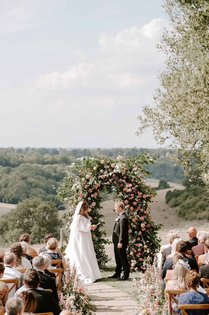 Bride in long sleeve wedding dress reading her vows to groom in black tux at outdoor wedding at Botley Hill Barn wedding venue 