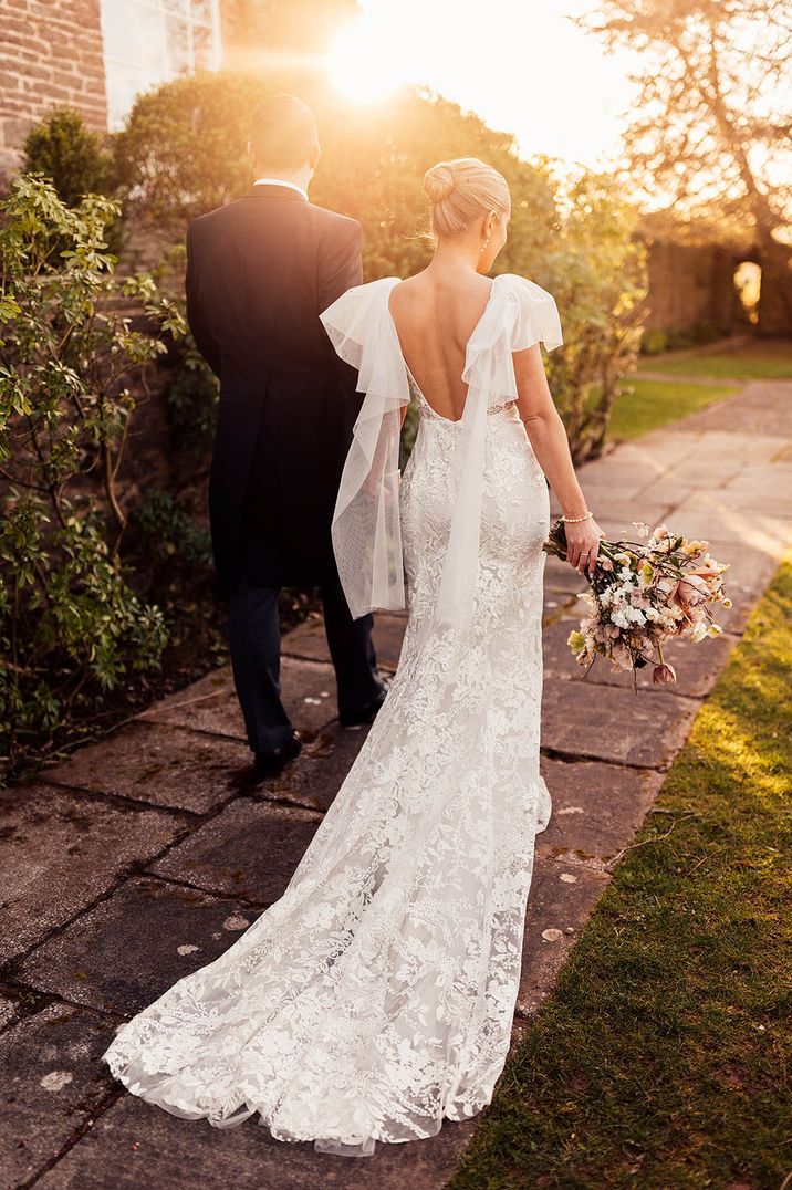 Bride in a low back lace wedding dress with tulle wings walking along with the groom during golden hour 