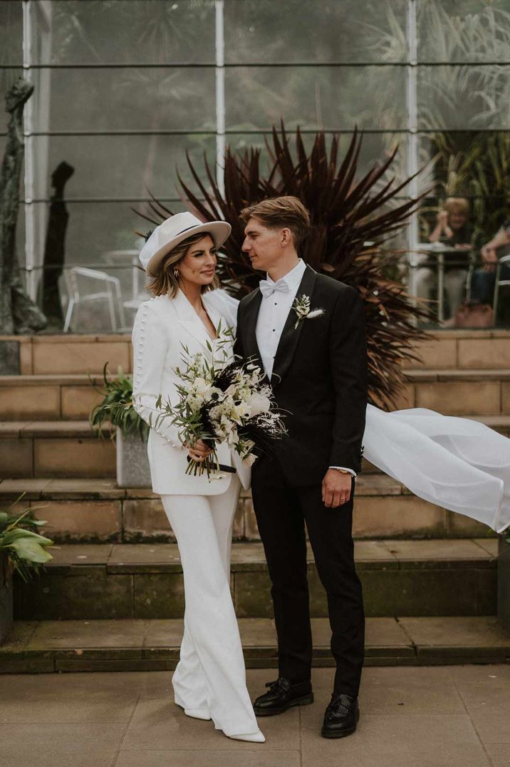 Bride in all white bridal suit, church length wedding veil and white wedding hat standing with groom in classic black tuxedo 