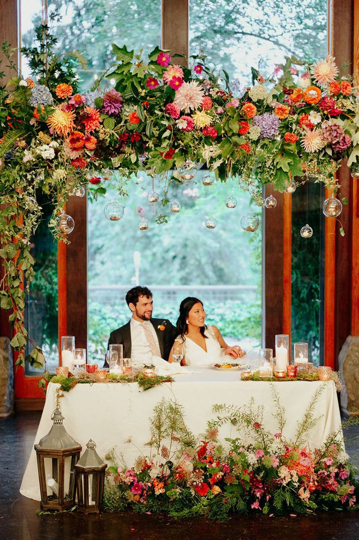 The bride and groom sit together under pretty flower arch at their sweetheart table 