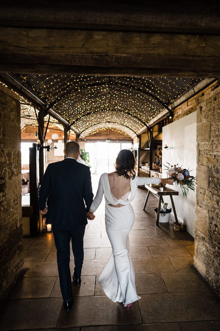 Bride in a cowl back wedding dress with lace insert walking through her rustic wedding venue with her husband 