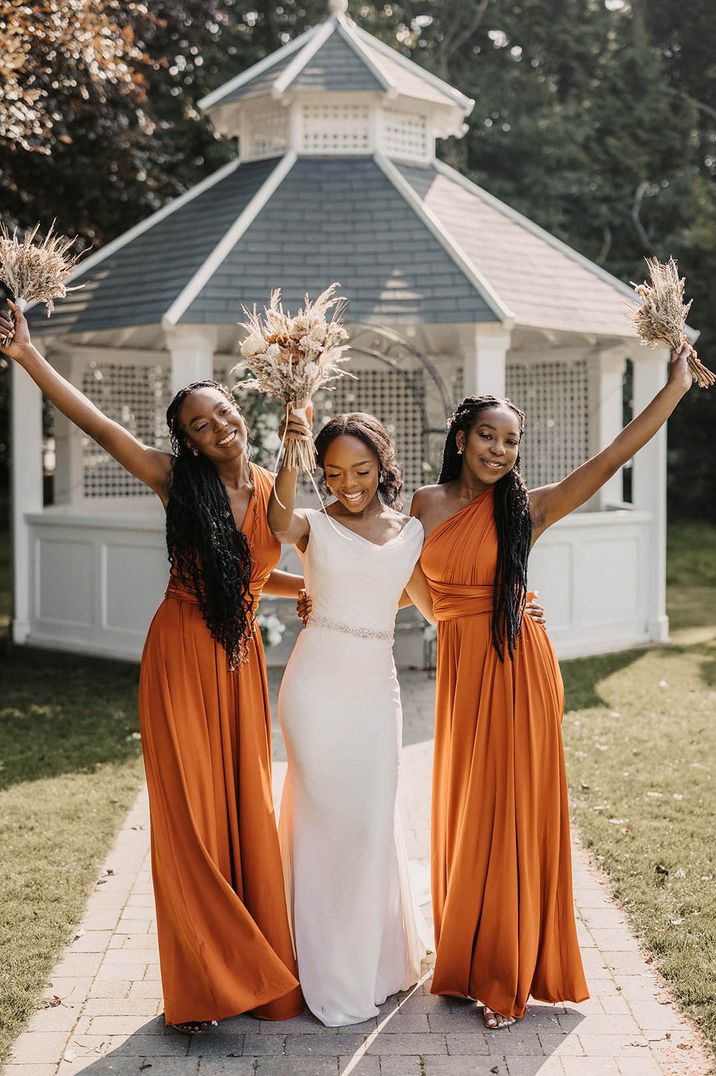 Bridesmaids in burnt orange satin bridesmaid dresses with the bride in an off the shoulder wedding dress smiling together 