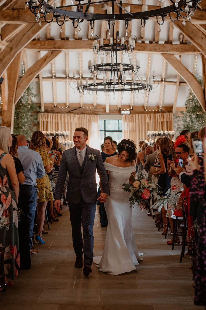 Bride in off the shoulder satin wedding dress and groom in dark suit walking down the aisle at Tithe Barn