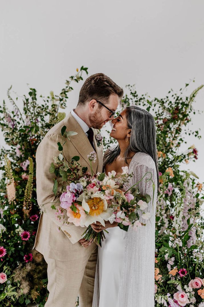 Groom in beige suit smiling down at the bride in a silver tassel bridal cape in front of colourful flower columns 