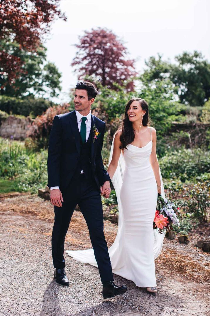 Bride in slight cowl neck strappy wedding dress walking hand in hand with groom in classic three piece black suit and boutonniere at Elmore Court - wedding venue search 