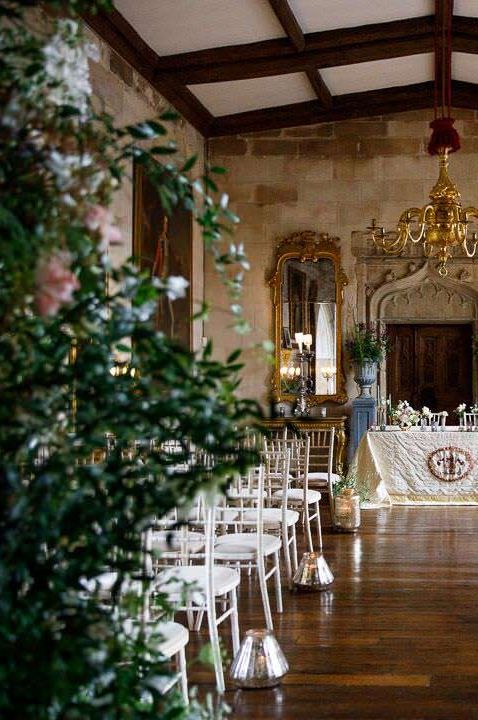 Ceremony room of Berkeley Castle wedding venue with large golden chandelier, wooden ceiling beams, foliage arrangements, large golden candle holder aisle decor and large golden framed mirrors