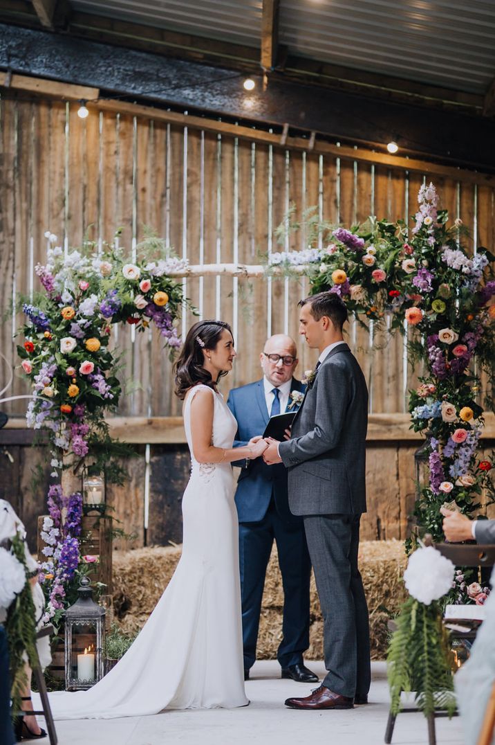 Bride in a fitted wedding dress standing at the colourful flower altar holding hands with her groom in a grey wedding suit 