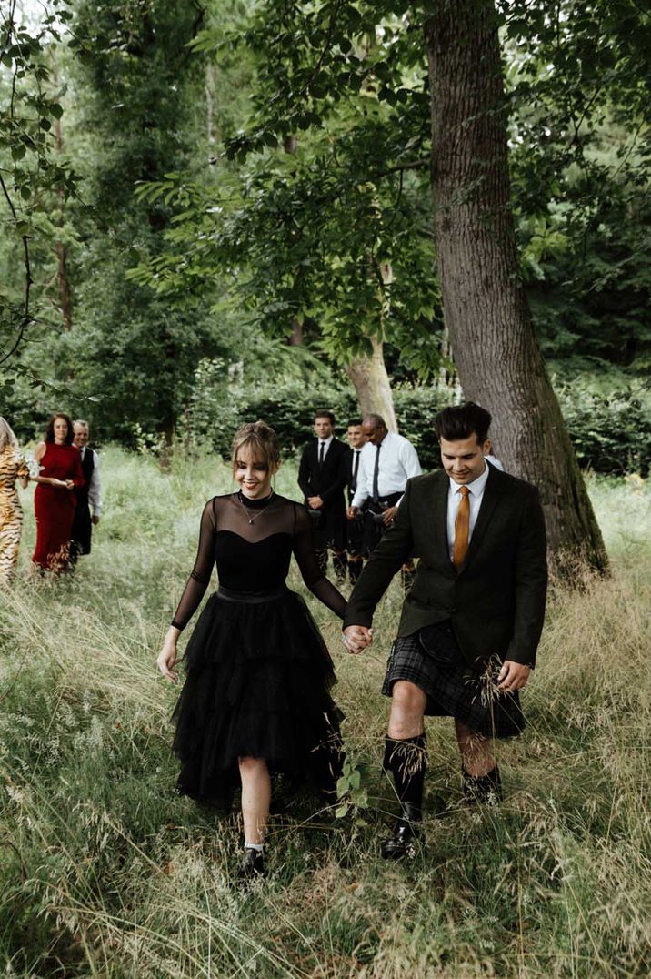 Bride in long sleeve black wedding dress with sheer neckline and sleeves holding hands with groom in black blazer and kilt 