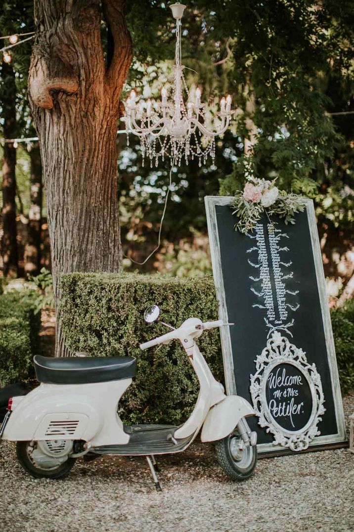 White wedding moped underneath a chandelier and next to large chalkboard wedding sign 