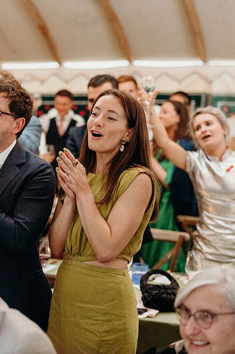 Wedding guests wearing lilac and olive dresses clapping and celebrating during wedding reception while drinking champagne