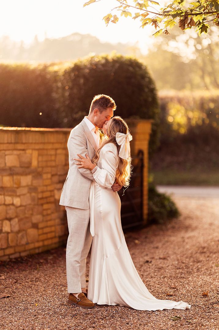 Bride in long sleeve satin wedding dress and hair bow accessory with the groom in a grey suit kissing during golden hour 